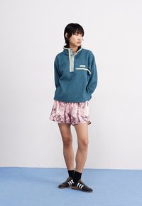 Tyrkysový fleecový pulóver s vysokým golierom, svetlomodrými akcentmi a zipsovým vreckom na hrudi. V páre s ružovými šortkami s vzorom a čiernou obuvou.