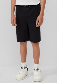 Zwarte shorts met een elastische tailleband en trekkoorddetail, met een rechte snit en een gladde stof. Draagt bij witte sneakers en sokken.