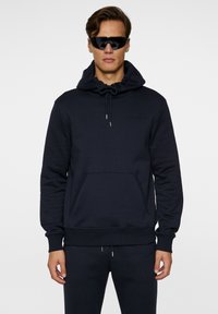 Mørk navy sweatshirt med snørehætte, frontlomme, blød struktur og diskret logo-detalje på brystet.