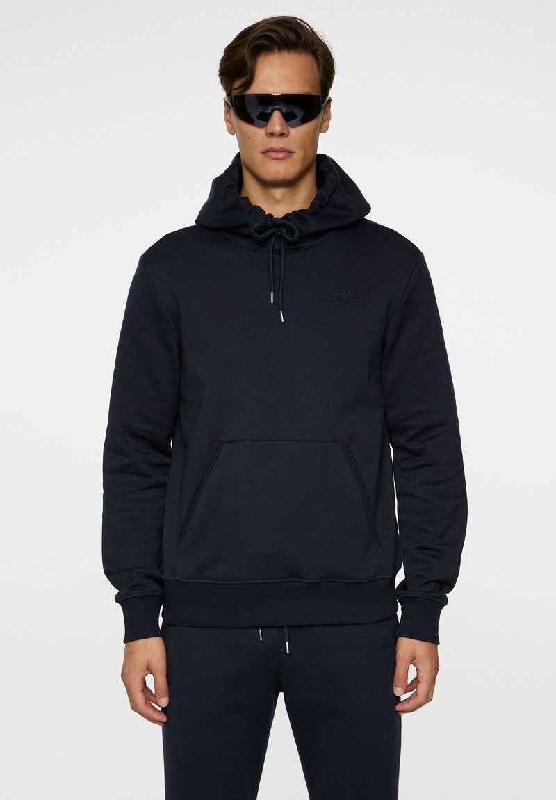Mørk navy sweatshirt med snørehætte, frontlomme, blød struktur og diskret logo-detalje på brystet.