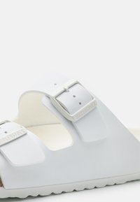 Baltas Birkenstock sandāls ar divām regulējamām siksnām ar sudraba sprādzēm, korķa pamatni un gumijas ārpusi ar reljefu.
