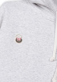 Sudadera gris con un acabado texturizado, que presenta un pequeño parche colorido de un sombrero con rayas rojas en el área del pecho izquierdo.