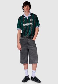 Grön och svart randig tröja med vit krage, grå denimshorts, vita strumpor och svarta skor. Tröjan har en logotyp och ett texturerat mönster.