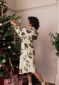 Hætteklædt badekåbe i cremefarvet fleece i knælængde med julemotiver, herunder leopardprint og kristtorn, båret under pyntning af et juletræ.