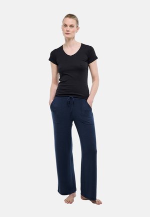 Femme debout pieds nus, mains dans les poches, portant un haut noir à manches courtes et un pantalon ample bleu marine avec un cordon de serrage.