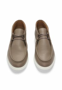 Scarpe in pelle marrone con superficie textured, punta rotonda e suole in gomma bianca. Presentano due fori per i lacci e cuciture visibili attorno ai bordi.
