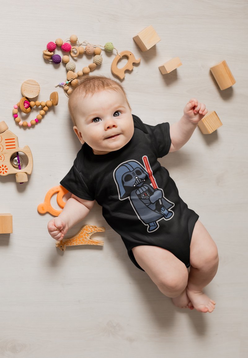 Černá overal s kresleným Darth Vaderem a červeným světelným mečem. Kolem se nacházejí dřevěné hračky v podobě bloků a korálků v různých barvách a strukturách.