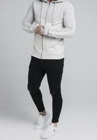 Uomo che indossa una felpa con zip grigia chiara, pantaloni neri attillati, sneaker bianche con dettagli dorati, mani tatuate e caviglia visibile.