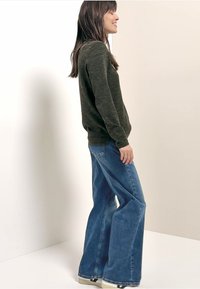 Tmavě zelený svetr s texturou v kombinaci se světle modrými rozšířenými džíny. Oblečení je jednoduché, s dlouhými rukávy a volným střihem.