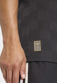 Black athletic shirt with textured pattern, yellow branded tag, and contrasting striped accents at hem. Material appears smooth and breathable.