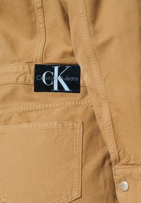Gros plan sur un tissu beige avec un patch logo Calvin Klein Jeans noir et blanc au-dessus d'une grande poche cousue et une manche retroussée avec un bouton métallique.