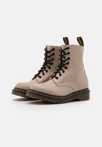 Botas de tobillo en color beige claro, hechas de cuero suave, con cordones negros, costuras amarillas y una suela de goma negra de diseño robusto.