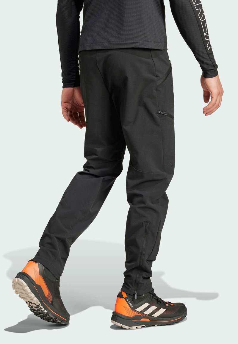 Zwarte outdoorbroeken hebben een slim fit, meerdere met rits gesloten zakken en een zachte, lichtgewicht textuur. Gedragen met zwart-oranje wandelschoenen.