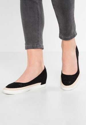 Czarne baleriny z wyciętym wzorem, biała podeszwa w stylu espadryli i teksturowany materiał. Noszone z szarymi spodniami 3/4 na białej powierzchni.