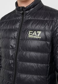 Giacca a vento nera con sezioni imbottite, colletto alto e cerniera frontale. Logo EA7 Emporio Armani in verde chiaro.