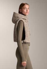 Fiatal nő bézs színű, ujjatlan fleece mellényben kapucnival, olíva zöld bordázott hosszú ujjú felső és hozzá illeszkedő leggings viselésével, oldalról áll.