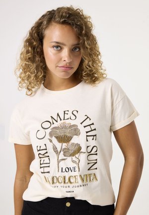 T-shirt en coton crème avec un design floral aux accents dorés et noirs et le texte "HERE COMES THE SUN" dans une police arrondie.