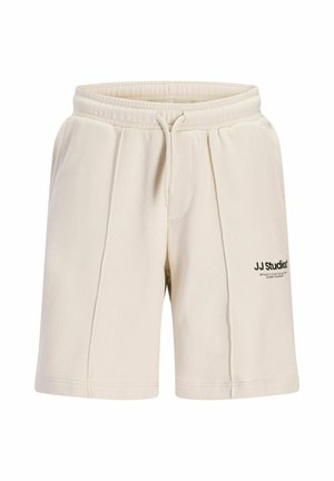 Nastolatek stoi, ubrany w czarną koszulkę z nadrukiem, kremowe szorty, białe skarpetki do kostek i białe trampki z czarnymi akcentami.