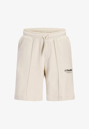 Nastolatek stoi, ubrany w czarną koszulkę z nadrukiem, kremowe szorty, białe skarpetki do kostek i białe trampki z czarnymi akcentami.