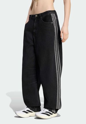 Persona che indossa pantaloni neri larghi con tre strisce laterali grigie e scarpe da ginnastica bianche con strisce nere, in piedi davanti a uno sfondo uniforme.