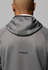 Szara kurtka z kapturem, wykonana z wzorzystego materiału, z czarnym logo na górnej części pleców, charakteryzująca się gładką teksturą i regulowanym kapturem.