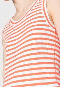 Débardeur rayé orange et blanc en tissu côtelé. Présente un col rond et une coupe ajustée. Couture accentuée sur les bords.