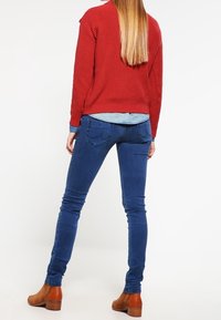 Pull rouge en tricot assorti à un jean skinny bleu, des bottines marron et une chemise chambray visible à la taille.