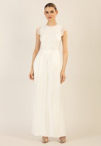 Weißer Jumpsuit mit einem Spitzentop und kurzen Puffärmeln, ausgestattet mit einer taillierten Taille und weiten, fließenden Hosenbeinen. Silberne Akzente an den Schuhen.