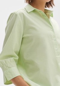 Camicia verde chiaro con colletto, maniche a tre quarti e polsini arricciati. Realizzata in tessuto morbido e strutturato.