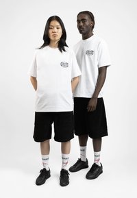 Bílé unisex trička, černé manšestrové kraťasy, černé tenisky a vzorované bílé ponožky. Tričko má malé černé grafické logo.