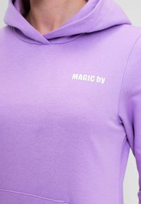 Felpa viola con tasca frontale e cappuccio regolabile con cordini. Presenta il testo bianco "MAGIC by" sul petto. Tessuto morbido in misto cotone.