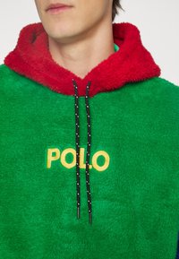 Měkká zelená mikina s červenou kapucí lemovanou kožešinou, s žlutým logem "POLO" a černými stahovacími šňůrkami s bílými tečkami.
