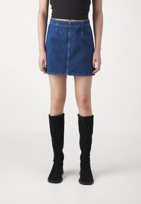 Kvinna iklädd en blå jeans minikjol, svart crop top och knähöga svarta stövlar, ståendes mot en plain vit bakgrund.