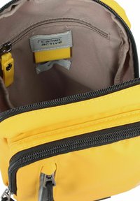 Mochila de tela amarilla con cierre de cremallera, que presenta un interior beige, etiqueta gris y una pequeña correa amarilla en su interior para organización.
