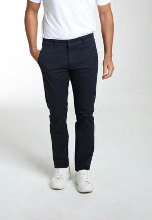 Homme portant un pantalon bleu marine foncé slim, des baskets blanches et un t-shirt blanc rentré, debout avec une main dans la poche.