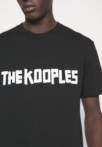 Svart bomull t-shirt med korta ärmar som har djärva, vita, utskurna bokstäver med texten "THE KOOPLES." Enkel rund halsringning, inga ytterligare mönster.