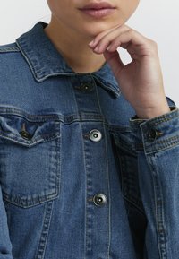 Modra jeans jakna s klasičnim dizajnom, črnimi gumbi, žepoma na prsih in vidnimi šivi na ovratniku in manžetah.