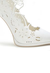 White high-heeled shoe featuring cut-out floral patterns, textured leather, and lace-up detail. Curved shape with a smooth sole.