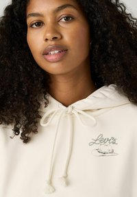 Cremfarbener Hoodie mit Kordelzugverschluss, ausgestattet mit einem kleinen, mehrfarbigen "Levi's"-Logo und weichem Stoffgefühl.