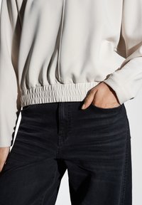 Persona che indossa una giacca elasticizzata di un beige chiaro con una mano nella tasca anteriore di jeans in denim neri.