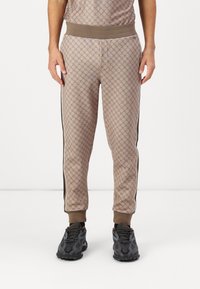Beige gemusterte Jogginghosen mit geometrischem Design, elastischem Bund und seitlichen Streifen. Kombiniert mit dunklen Sportschuhen.