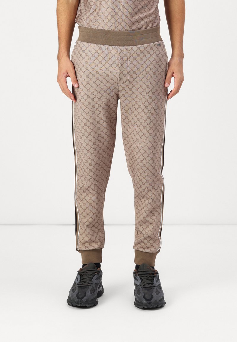 Beige gemusterte Jogginghosen mit geometrischem Design, elastischem Bund und seitlichen Streifen. Kombiniert mit dunklen Sportschuhen.