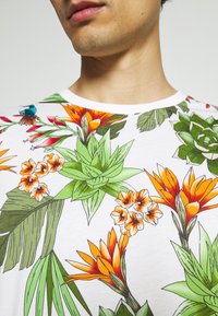 Camiseta blanca de manga corta con vibrantes estampados de flores y hojas tropicales en verde, naranja, amarillo y rojo. Tejido de algodón suave.