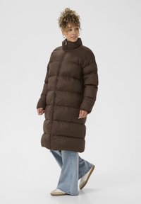 Hnedá puffová bunda s vysokým golierom, ktorá má horizontálne prešívané sekcie, predný zips a voľný strih, oblečená cez svetlomodré nohavice s rozšírením.