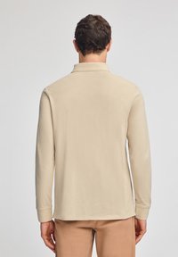 Beżowa koszulka polo z długim rękawem, z kołnierzykiem i prostym dołem, wykonana z miękkiej tkaniny. Plecy modela widoczne na neutralnym tle.