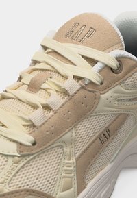 Μπεζ και κρεμ αθλητικό παπούτσι με ύφασμα διχτυωτό, λεπτομέρειες από σουέντ, κρεμ κορδόνια και το λογότυπο της GAP στη γλώσσα και στην πλαϊνή πλευρά.