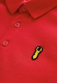 Polo rosso a maniche corte, con una trama texturizzata e due bottoni. Logo di un'aragosta gialla ricamato sul lato sinistro del petto.