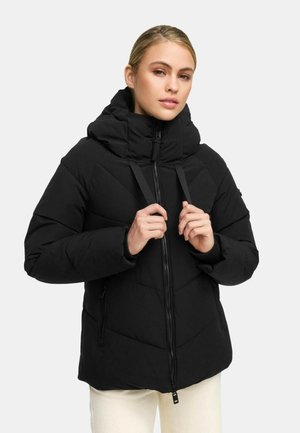 Woman adjusting the hood strings of a black quilted winter jacket with zipper, wearing light-colored pants, against a plain background.