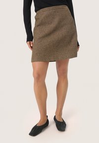 Jupe en tweed marron avec une taille cintrée et une forme évasée, présentant une texture subtile et un ourlet au-dessus du genou. Portée avec des ballerines noires.