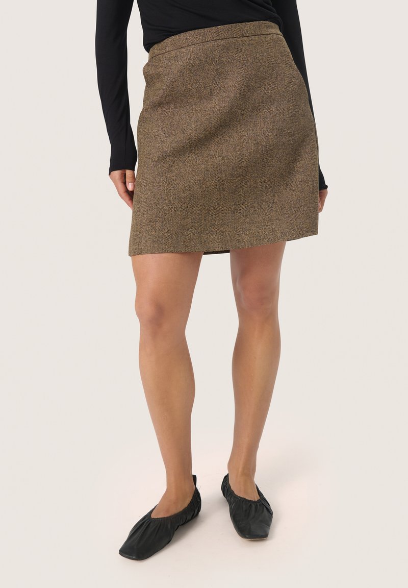 Jupe en tweed marron avec une taille cintrée et une forme évasée, présentant une texture subtile et un ourlet au-dessus du genou. Portée avec des ballerines noires.